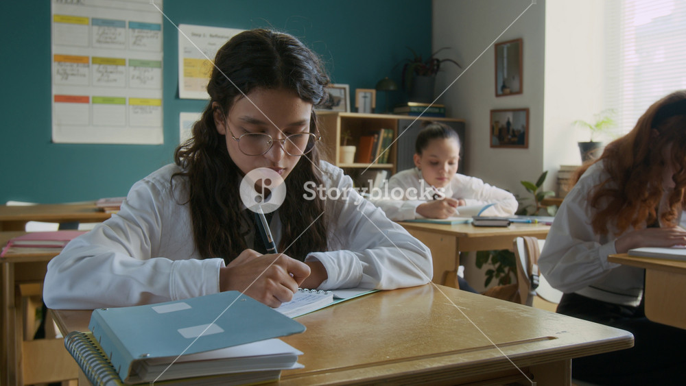 Medium portrait of focused teen Caucasian student girl in uniform writing in exercise book then looking at camera sitting at desk in well-equipped classroom studying in diverse single gender school