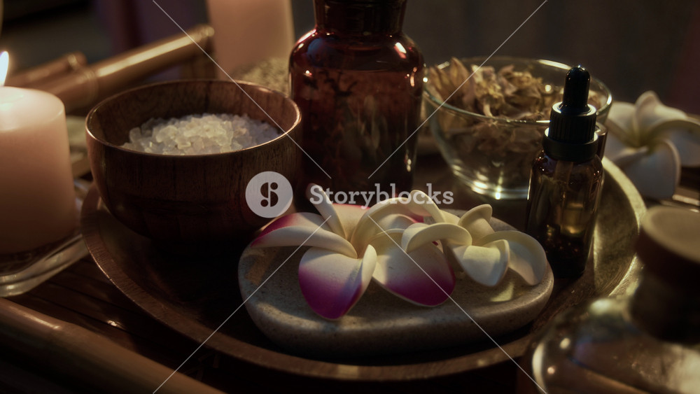 Close-up shot of wooden tray with coarse salt, plumeria blossoms, dried herbs, and essential oil bottle in massage salon