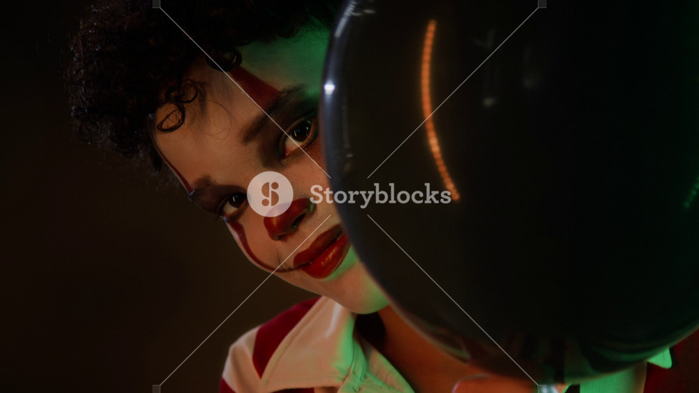 Portrait of teenage boy wearing creepy clown makeup, peeking from behind black balloon and smiling. Clip is perfect for Halloween marketing campaigns and horror movie trailers