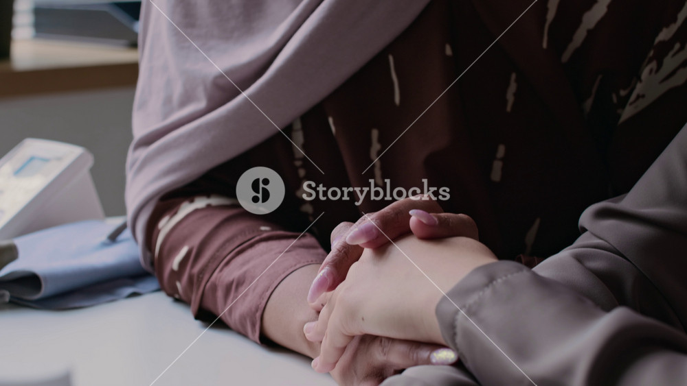 Tilt-up close-up shot of Muslim mother in hijab holding hands, supporting her daughter at medical appointment while talking to doctor in hospital