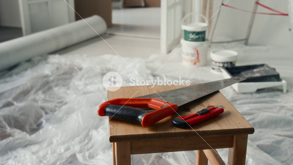Close-up view of handsaw with plastic handle and paper cutting knife on wooden stool in room undergoing renovation with paint buckets, tray and protective film covering floor