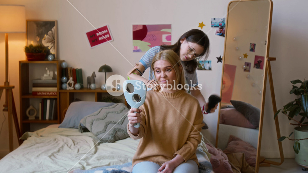 Teenage girl with prosthetic arm brushing long hair of her mother, holding hand mirror while sitting on bed in cozy room