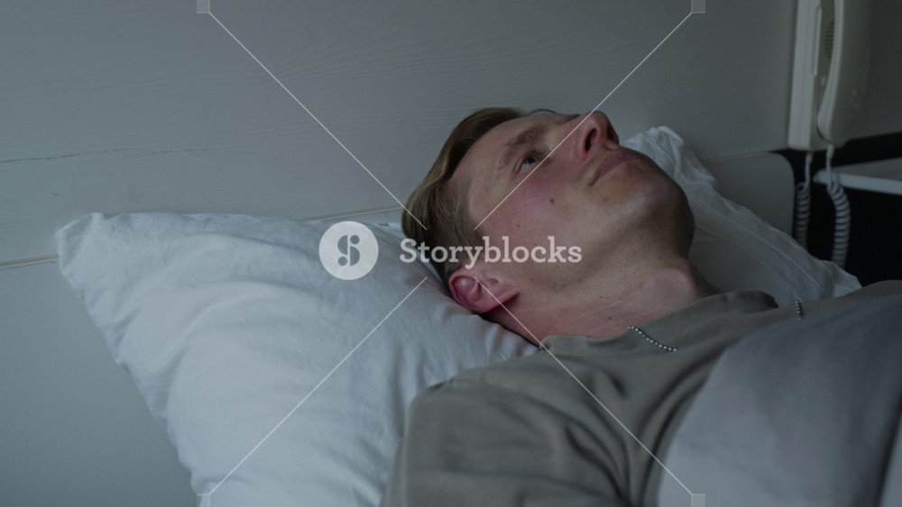 Young adult Caucasian man lying in bed awake, staring at ceiling with worried expression, suffering from insomnia. Video for mental health advocacy or sleep disorder treatment promotions