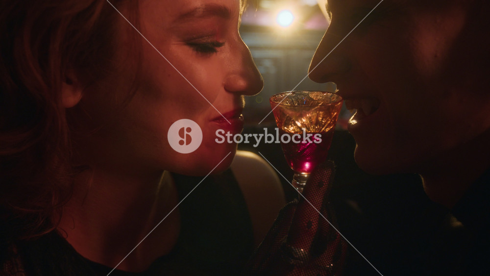 Close-up shot of smiling vampire woman offering glass of blood to man in intimate manner in dimly lit room warmed by candlelight