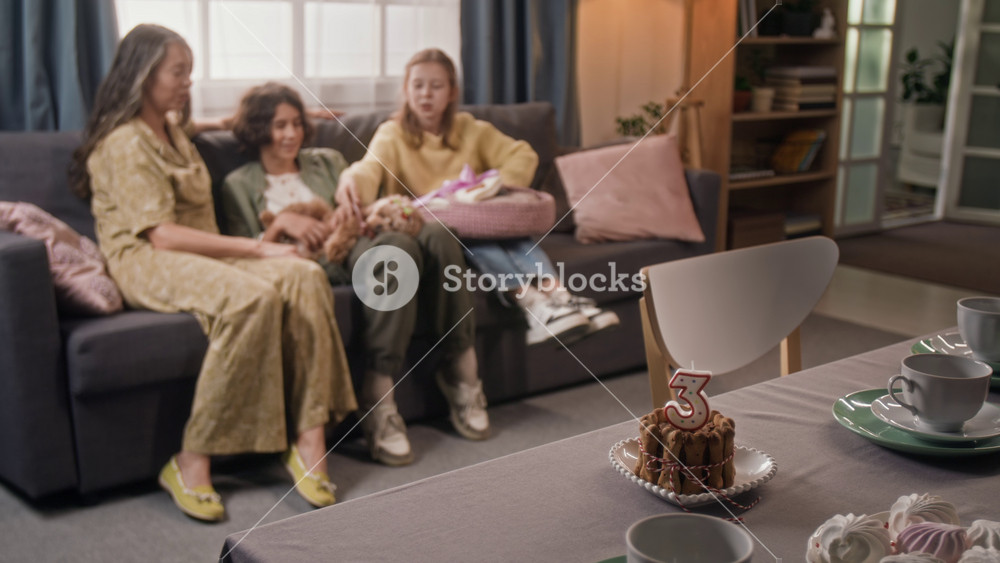 Rack focus shot of family of mother, two kids and maltipoo dog chatting and relaxing on couch in living room, table set with pet cake and cups