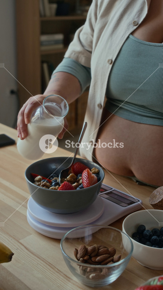 Vertical shot of unrecognizable woman pouring milk into bowl with granola and berries to have healthy breakfast at home