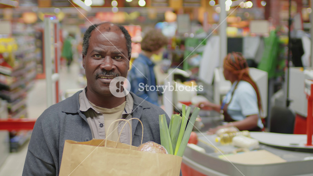 Shallow focus portrait of senior Black man holding paper bag full of food products standing in supermarket looking at camera