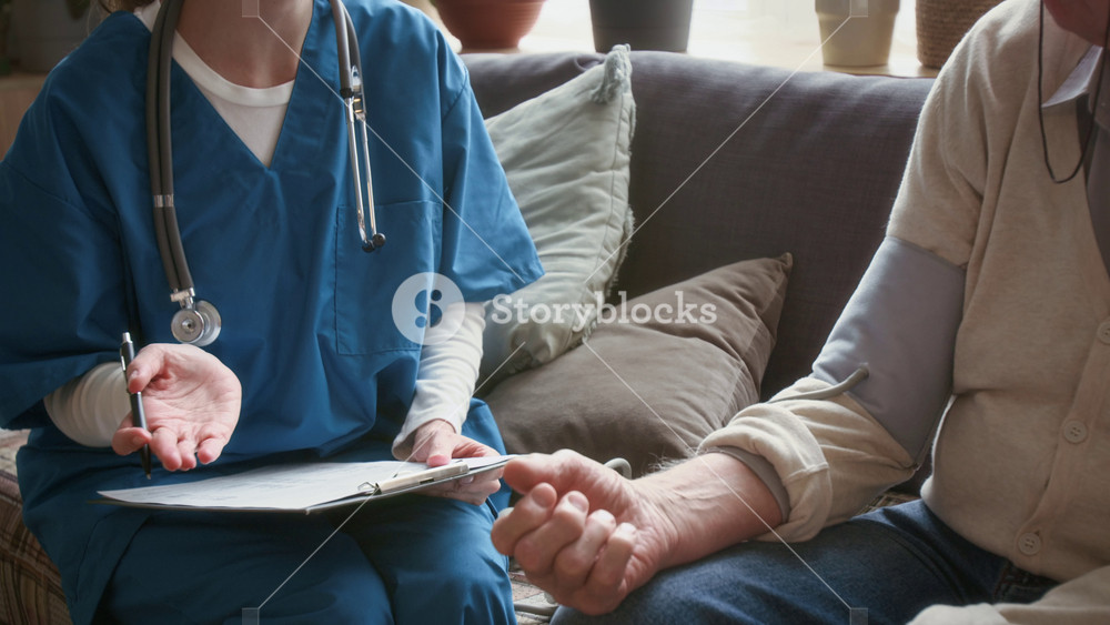 Tilt up shot of female doctor in blue scrubs consulting elderly patient and taking notes while measuring his blood pressure on couch in cozy living room of nursing home