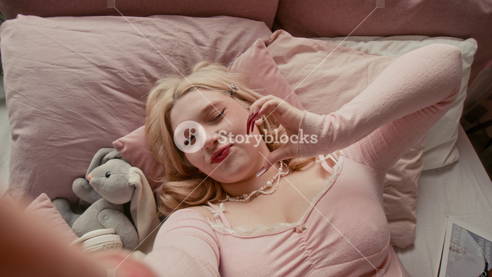 Portrait shot of smiling blonde girl in a pink lace top with long red nails holding camera and showing heart signs while lying in bed