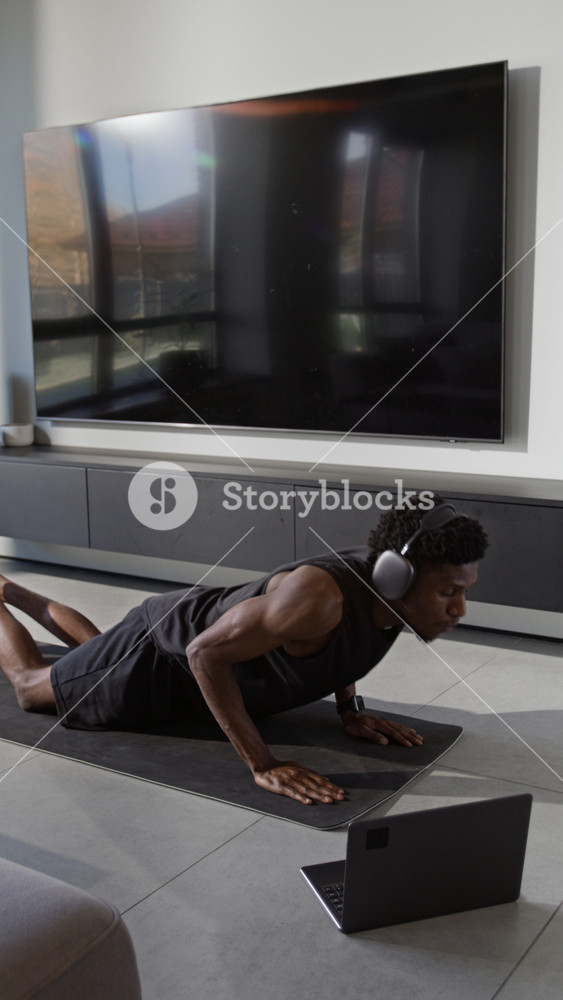 Vertical view of Black young man in sportswear with headphones lifting chest off fitness mat while performing cobra stretch pose during online yoga session
