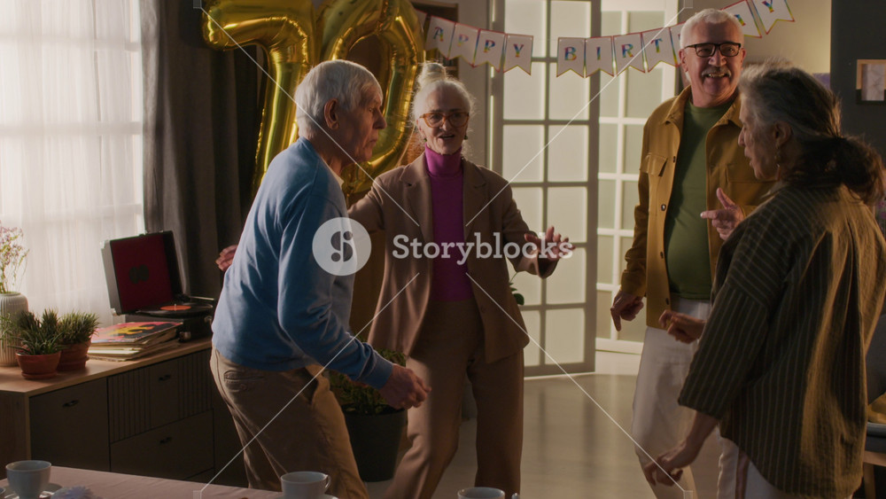 Company of four cheerful senior friends energetically dancing during birthday party of 70-year-old lady wearing pantsuit