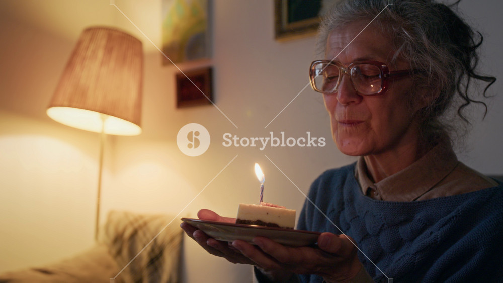 Happy elderly Caucasian woman with gray hair in bun and large glasses blowing candle on small piece of birthday cake