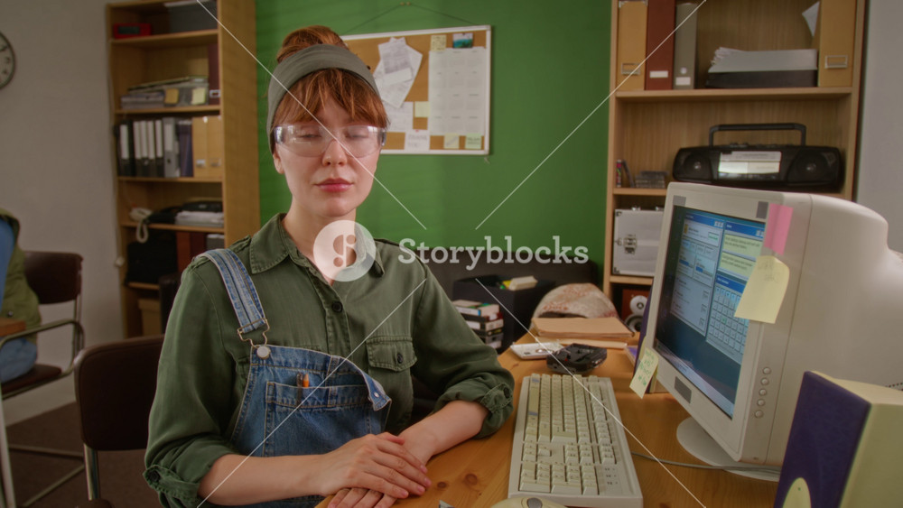 Portrait of Caucasian young adult female worker wearing safety glasses sitting at retro office desk next to vintage CRT computer, looking at camera. Ideal for retro tech marketing campaigns