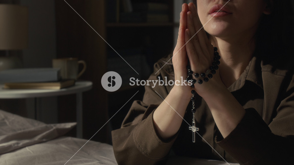 Lower face of gen Z girl dressed in brown shirt praying with hands pressed together holding black rosary beads with silver cross, then putting head down with closed eyes