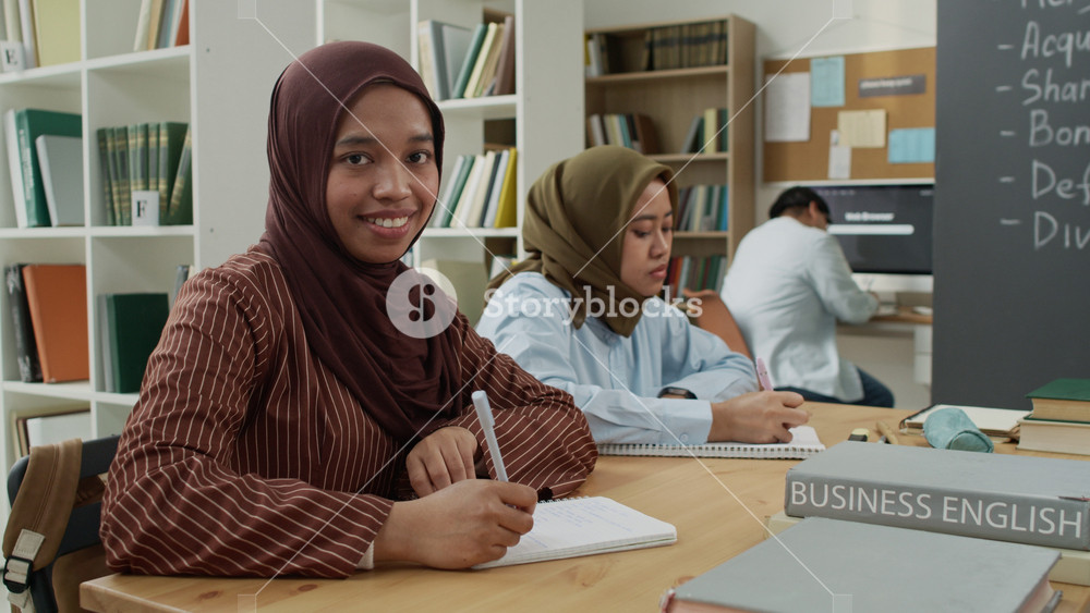 Portrait of Asian Muslim young adult female wearing hijab smiling at camera while studying in classroom. Footage is ideal for promoting inclusive education and diverse student representation