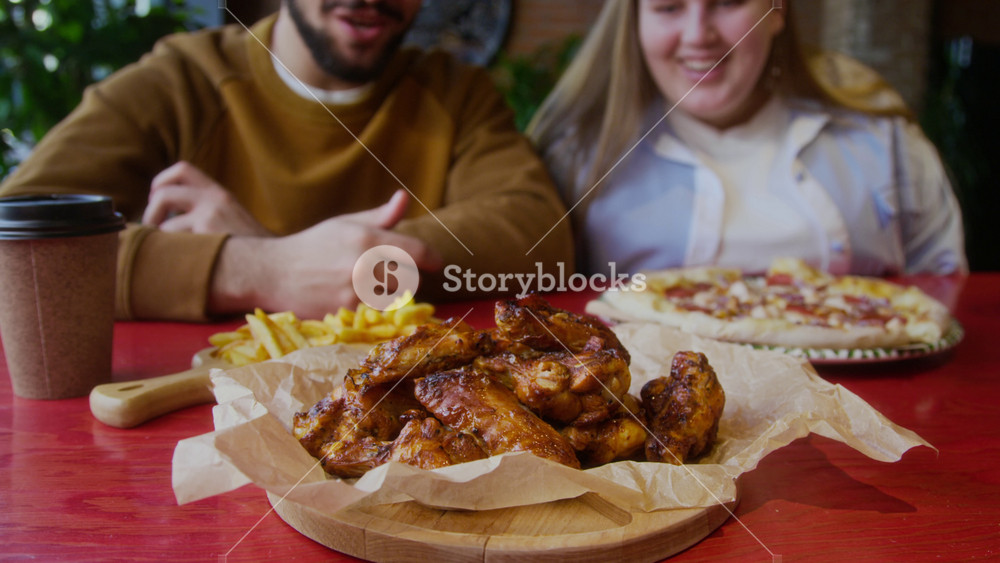 Tilt up shot of young man and smiling woman sitting at red table, surrounded by generous spread of mouthwatering baked chicken wings, pizza, french fries, and drink in cozy cafe