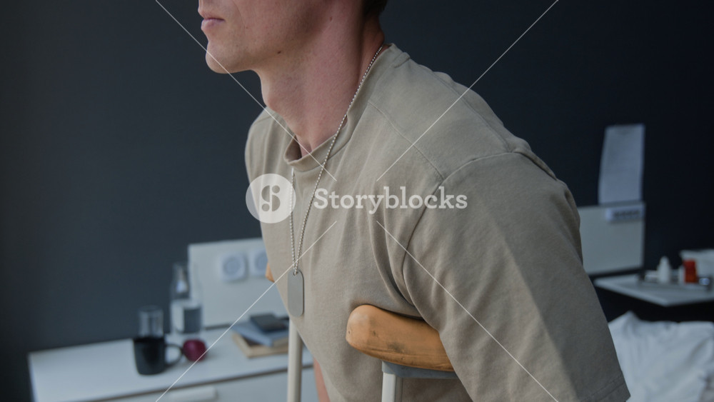 Tilt up shot of young Caucasian man with bandaged arm standing on crutches, looking forward. Footage for medical insurance, veteran rehabilitation, or healthcare recovery advertisements
