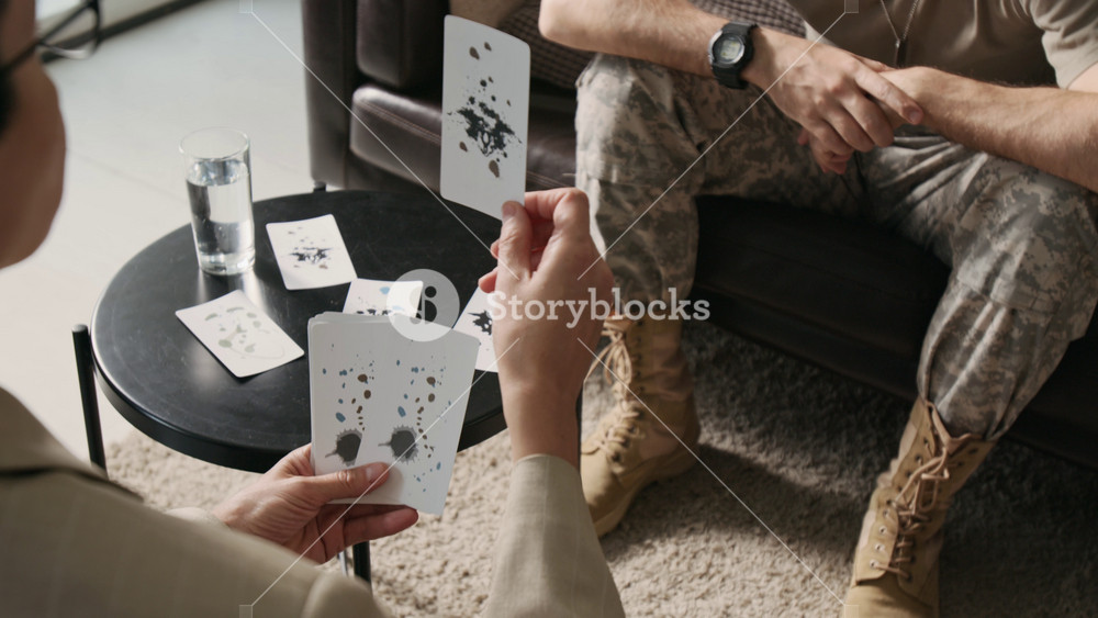 Over the shoulder shot of brunette female therapist holding cards with inkblots while carrying out psychological test for young veteran soldier