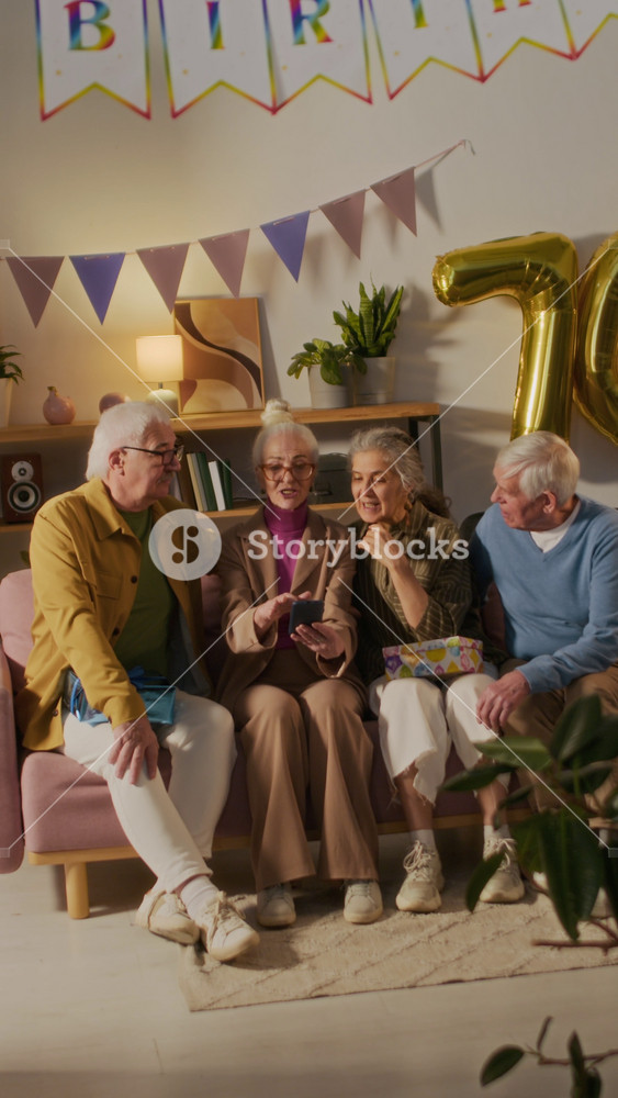 Vertical view of positive birthday lady showing videos on smartphone to three senior guests while sitting on couch all together in apartment decorated with flags and balloons