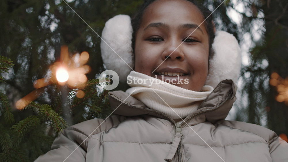 Close-up rack focus portrait of smiling African American gen alpha girl with earmuffs holding two burning sparklers in winter forest
