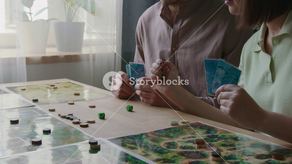 Tilt up shot of young boyfriend and girlfriend holding cards while playing strategic board game with game pieces and maps spread across table in living room