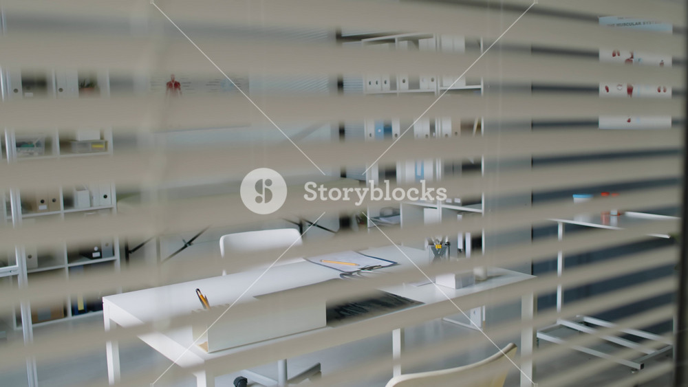 Tilt-down shot through blinds of modern, minimalistic medical office space with stationary and stethoscope on white desk, chair, examination couch and shelves with binders and posters in background