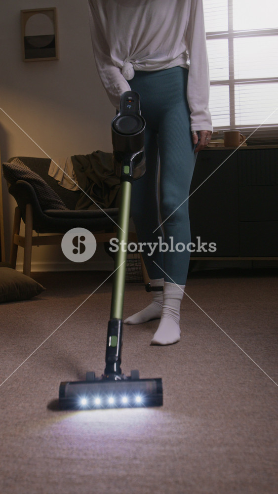 Vertical shot of unrecognizable woman cleaning her carpet with vacuum cleaner while wearing house arrest ankle monitor