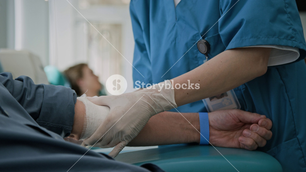 Close-up shot of unrecognizable nurse, wearing gloves and scrubs, applying bandage to arm of man after donating blood in clinic