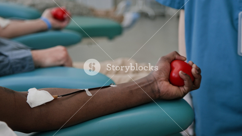 Close-up shot of unrecognizable healthcare worker assisting African American donor, squeezing red stress ball, covering insertion point in clinic