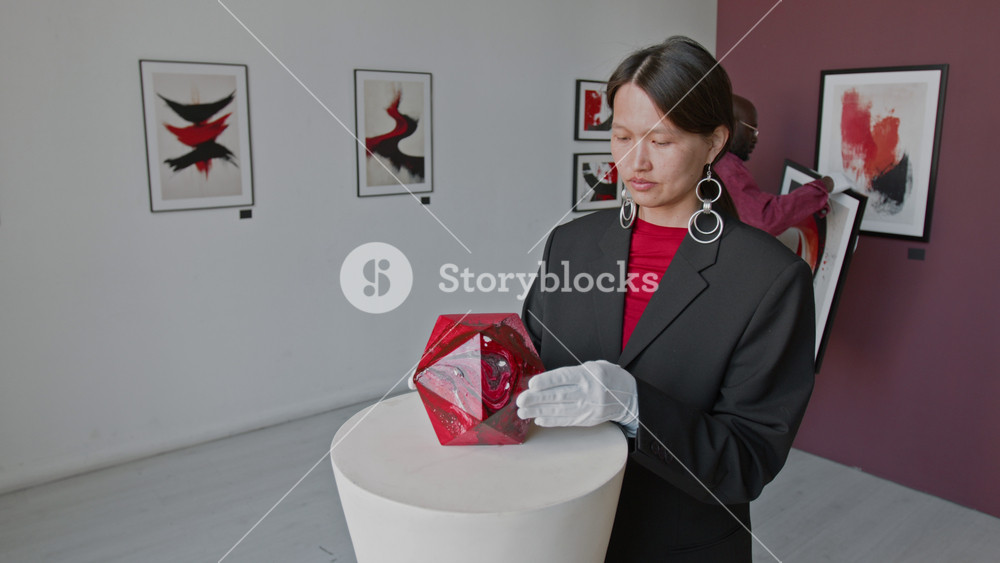 Asian woman in casual suit, wearing white gloves, putting multifaceted abstract red installation on pedestal, while Black man hanging painting on wall in art gallery