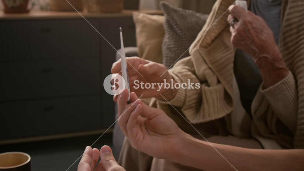 Tilt up shot of teenage boy taking electronic thermometer out of plastic case and handing it to sick senior woman, then he comforting granny while she measuring body temperature