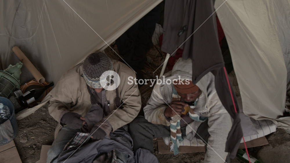 High angle tilt up view of Caucasian hobo sewing up holes in his pants while African American friend playing mouth organ, both sitting near tent outdoors