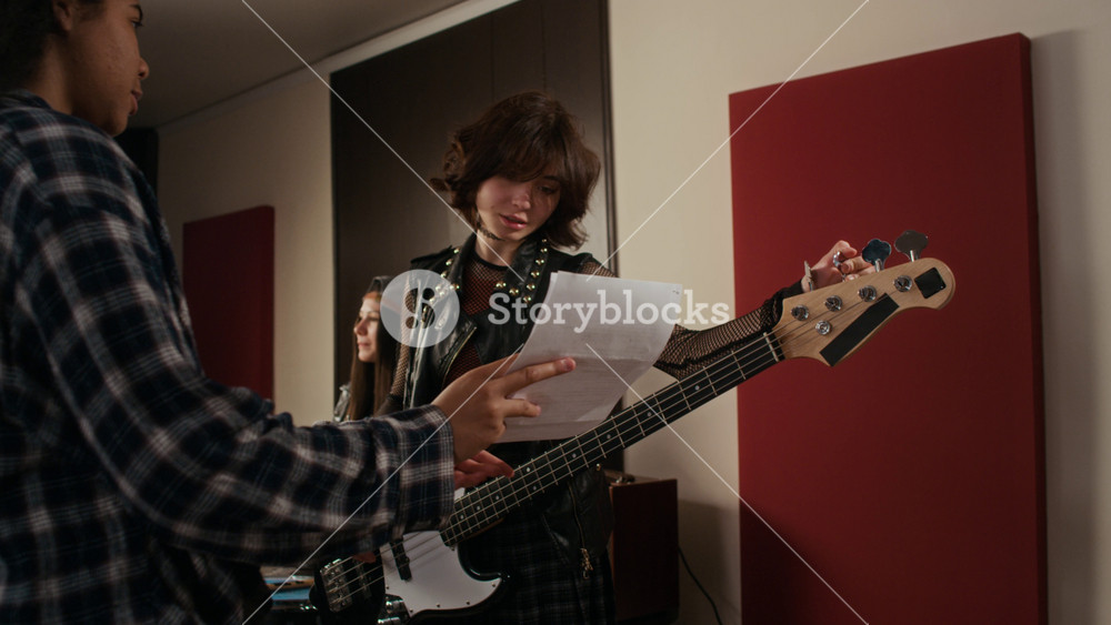 African American teenager showing sheet music to Caucasian teenager tuning bass guitar during rehearsal in studio. Video is ideal for youth culture, music education, and creative collaboration ads