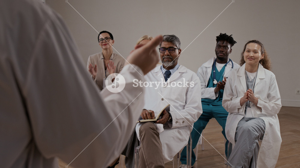 Unknown male doctor in lab gown referring to multiethnic audience including young and mature health workers and receiving applause from them after finishing his report at medical convention