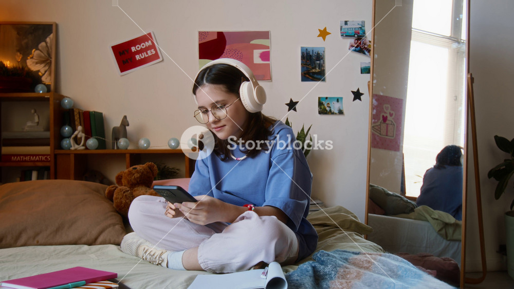Handheld zoom in shot of teenage girl listening to music in headphones while using her smartphone on bed in her room