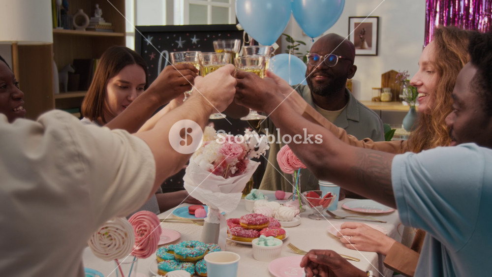 Over the shoulder view of company of multiethnic friends including young future parents clinking glasses with lemonade sitting at table set with sweet treats during gender reveal party