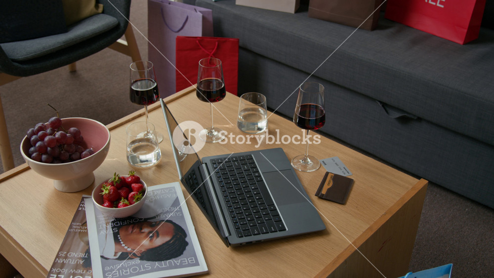 No people high angle shot of cozy living room with wooden coffee table laden with bowl of fruits, glasses of wine, magazines, and laptop