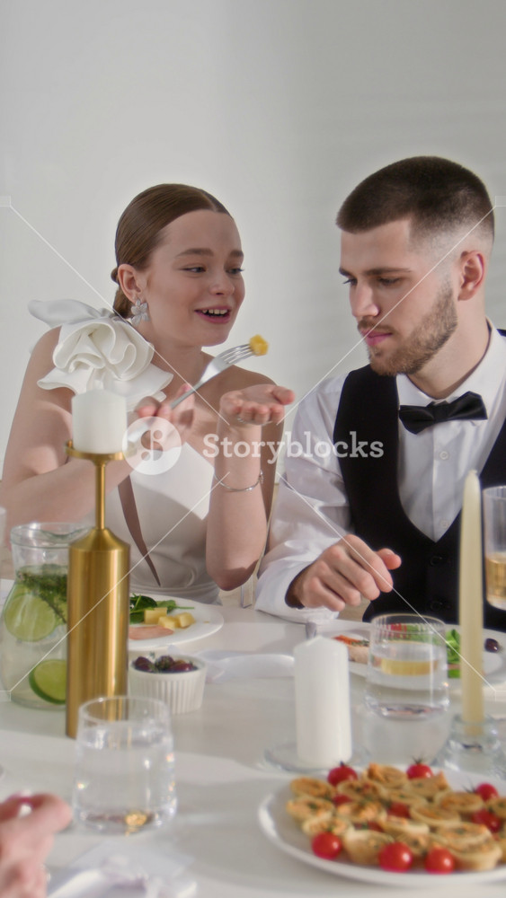Vertical shot of adorable newlywed couple in elegant dress and suit sitting at festive table and enjoying delicious food