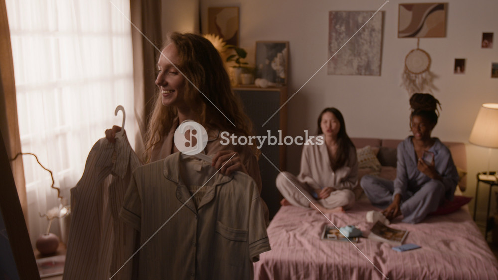 Medium shot of happy young Caucasian girl standing by mirror in bedroom, admiring new purchases, then asking diverse female friends for opinion, during fun pajama party at home