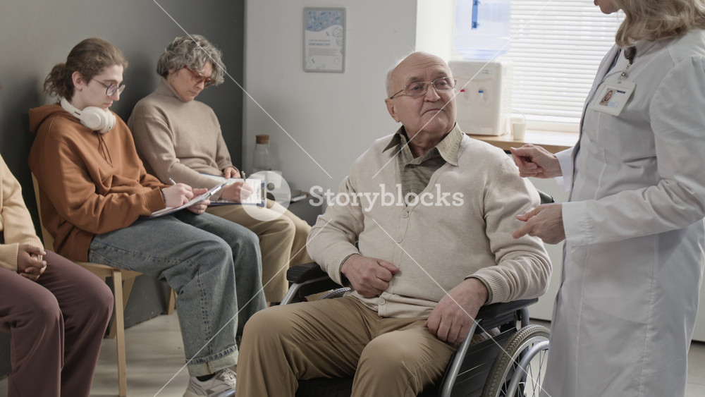 Mature Caucasian doctor working with senior man with disability in wheelchair, discussing his treatment and assisting, diverse patients sitting in background in waiting area