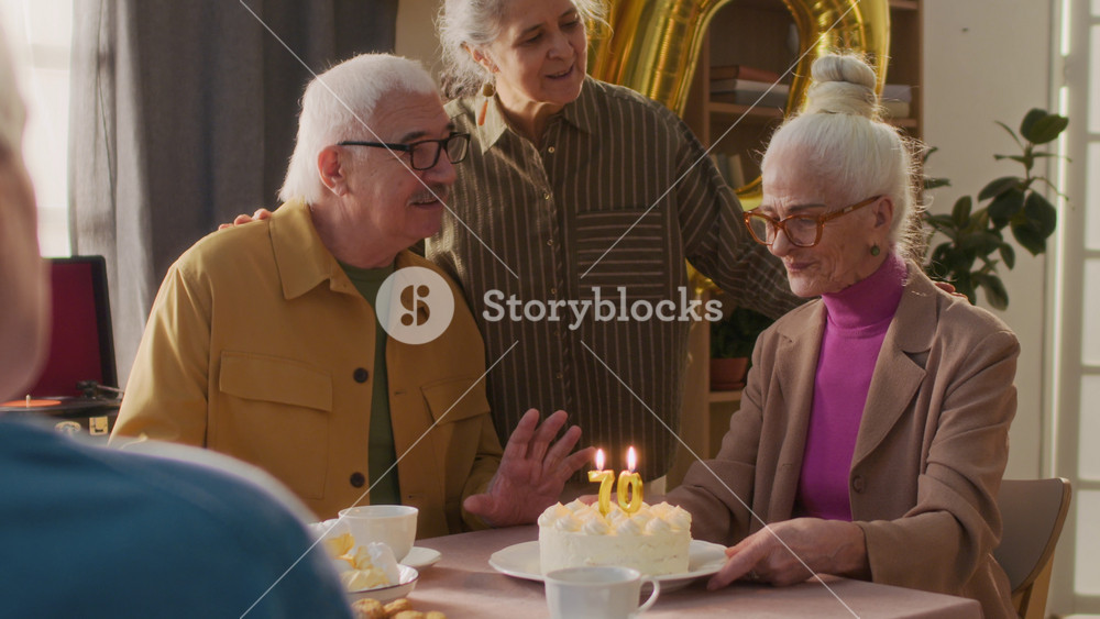 Over the shoulder shot of senior friends watching birthday lady making wish and blowing out candles sitting in front of cake at holiday table, all joyfully clapping hands