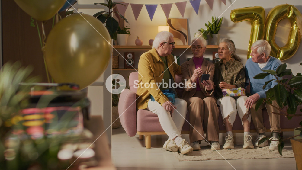 Senior woman showing digital photos on smartphone to three friends while sitting on couch together in living room decorated with balloons and party flags
