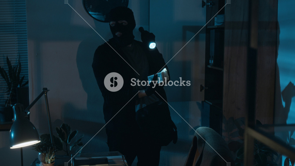 Rack focus view of young thief in balaclava finding money in wooden cabinet, he handing stack of dollar bills to his accomplice who smelling it and putting in bag