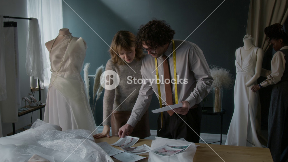 Modern bridal tailor consulting young Caucasian woman, showing pictures of wedding dresses to her, Black woman arranging dresses in background