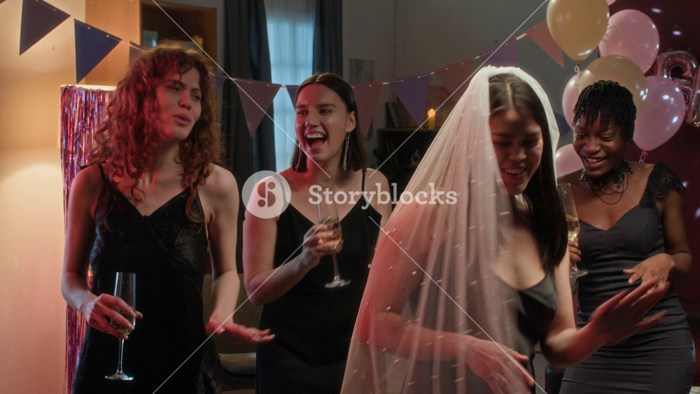 Group of young women in elegant dresses and bride-to-be in veil holding champagne glasses and dancing during festive bachelorette party in decorated room