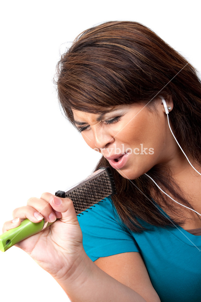 A Hispanic woman listening to music playing through her stereo earbud headphones and pretending to sing on a microphone that is actually a hair brush.