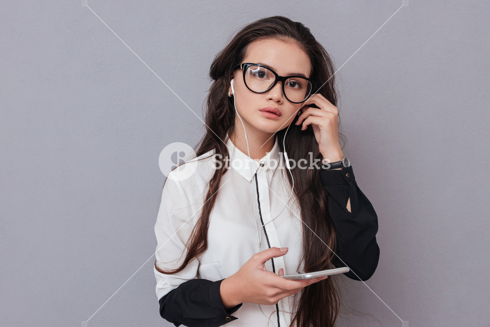 Attractive woman in glasses and shirt listening to music on phone. Isolated gray background.