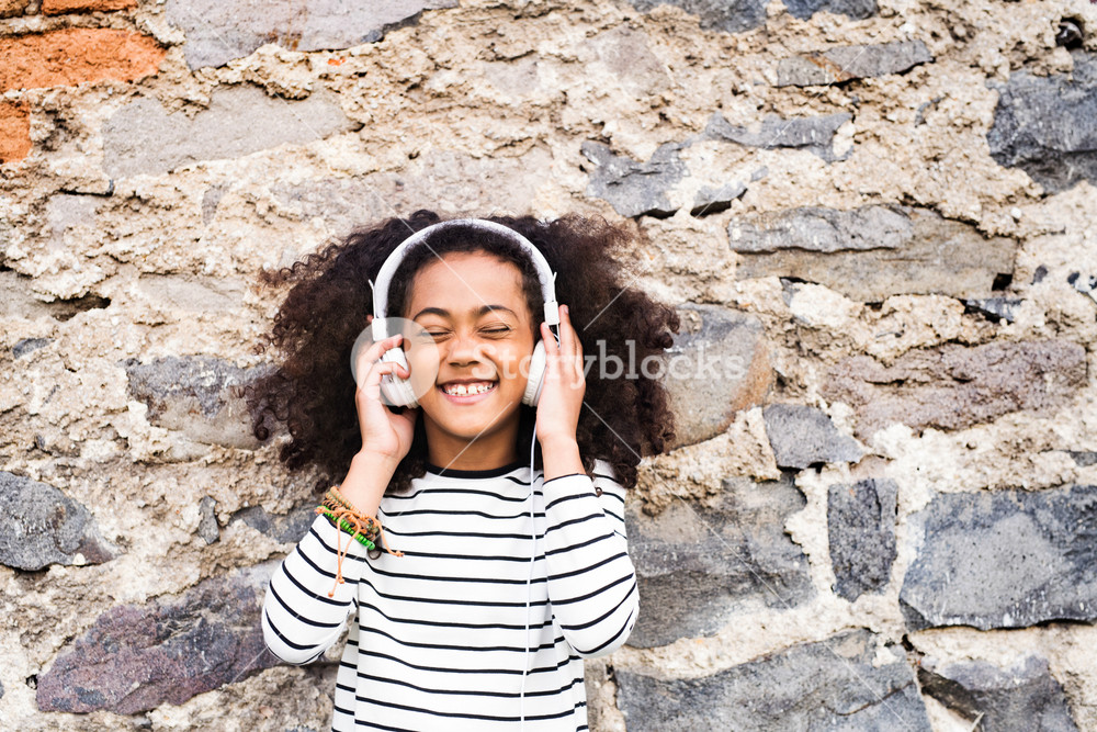 Stunning girl with curly hair outdoors, against stone wall, enjoying music.