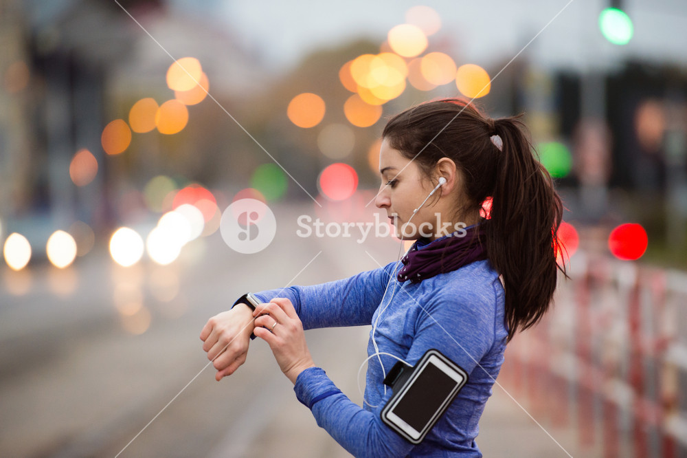 Young woman in the city with phone, watch, and earphones, enjoying music. Using a fitness app for progress tracking, running goals, or run summary. Rear view.