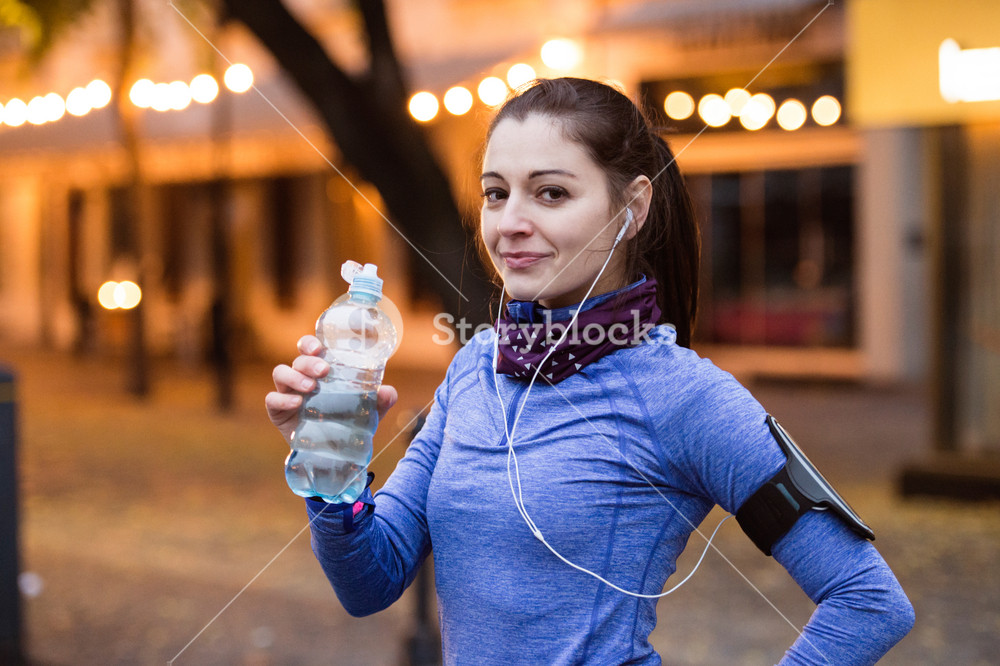 Young woman with smartphone and earphones, enjoying music, taking a break, hydrating. Jogging in the lit city at night.